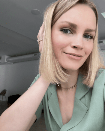 A person with shoulder-length blonde hair and blue eyes, wearing a green top, poses for a selfie indoors.