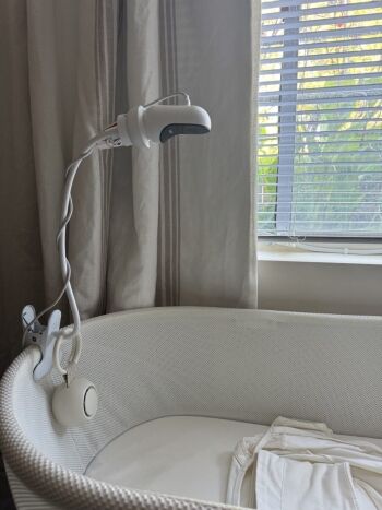 A white baby monitor camera is clipped to the side of a bassinet near a window with blinds and beige curtains, while massage guns rest neatly on a nearby table.