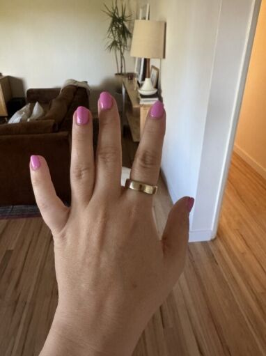A hand with pink nail polish and a gold oura ring is shown in the foreground, with a living room and houseplant visible in the background.