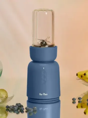Blue portable blender with a transparent blending cup on top, displayed upright on a reflective surface alongside massage guns, blueberries, and a banana.