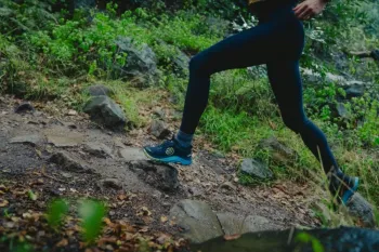 Person in black leggings and blue trail running shoes jogging uphill on a rocky, damp forest path surrounded by greenery.