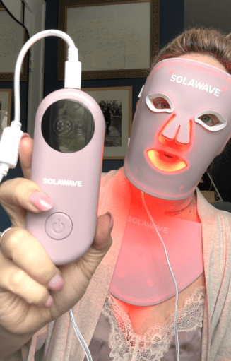 Person wearing a pink LED facial mask and neck device, holding a matching pink Solawave handheld controller, with both devices glowing red.