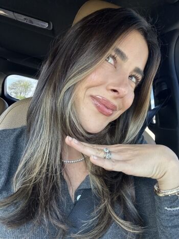 A woman with long hair smiles while sitting in a car. She rests her chin on her hand, displaying rings, and wears a bracelet and a necklace.