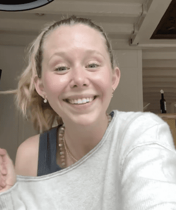 A smiling woman with blonde hair in a ponytail wearing a light sweater and necklace poses indoors.
