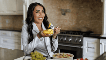 A woman sits in a kitchen, smiling and holding a bowl of fruit with a fork. Various fruits are on the counter in front of her.