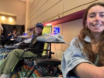 Two women sit in reclining chairs receiving oxygen therapy through nasal tubes in a modern, brightly lit room. Nearby, massage guns rest on a table, adding to the relaxing atmosphere as one woman smiles at the camera and the other looks on and smiles.