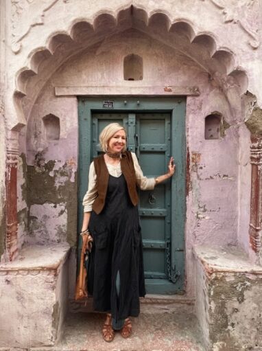 A woman stands smiling in front of a blue door set in a weathered pink wall with ornate architectural details.